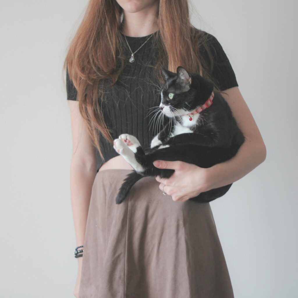 Young woman holding a tuxedo cat indoors, showcasing a minimalist fashion style.
